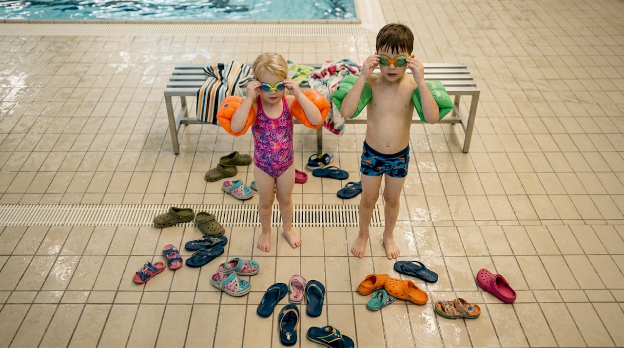 Kinder planschen mit Schwimmflügeln und Poolnudeln im Wasser, während ihre Eltern entspannt am Beckenrand zusehen.